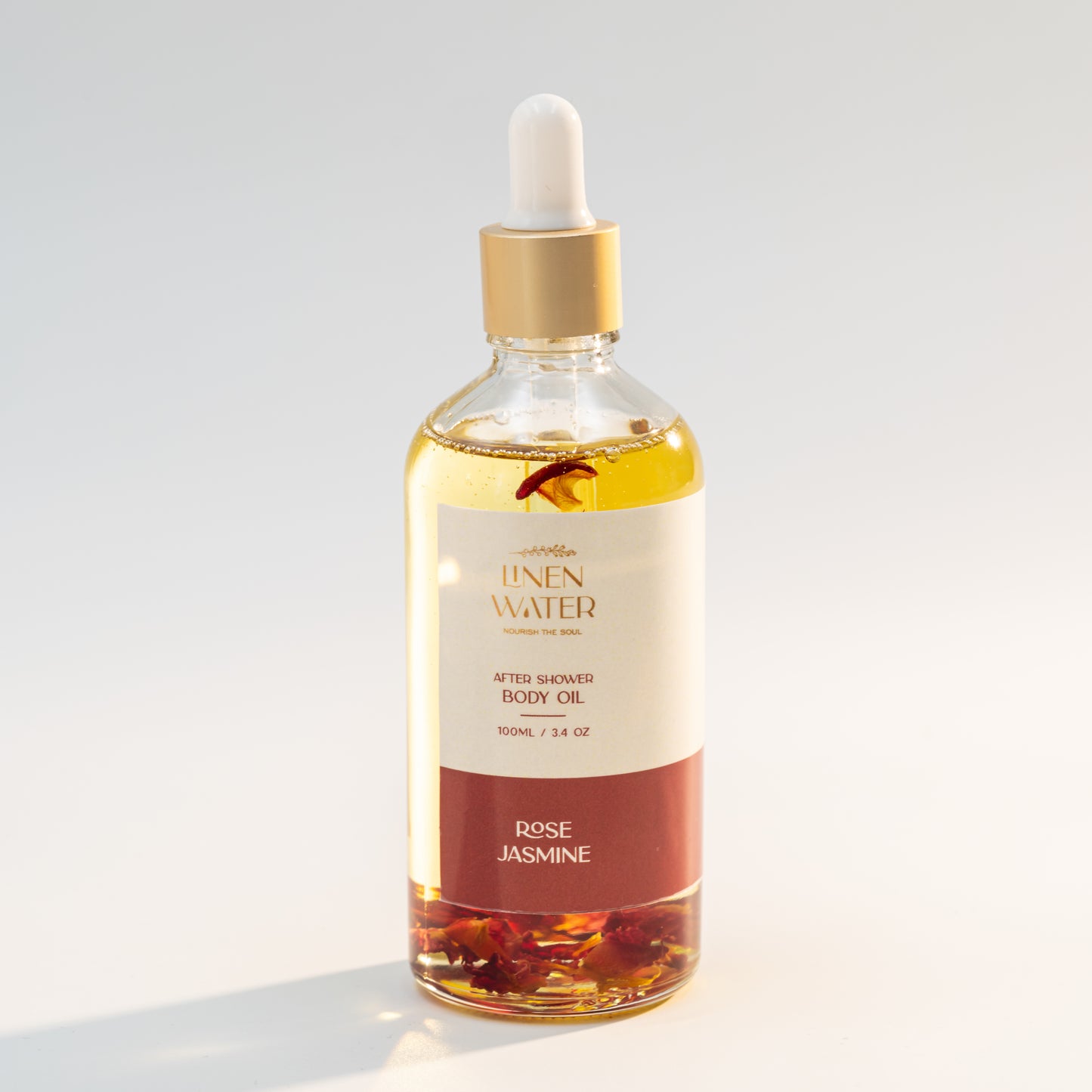 Fresh rose petals rest exquisitely inside a glass dropper bottle of Linen Water Rose Jasmine Body Oil, creating a clean, pure image when photographed against a stark white background.