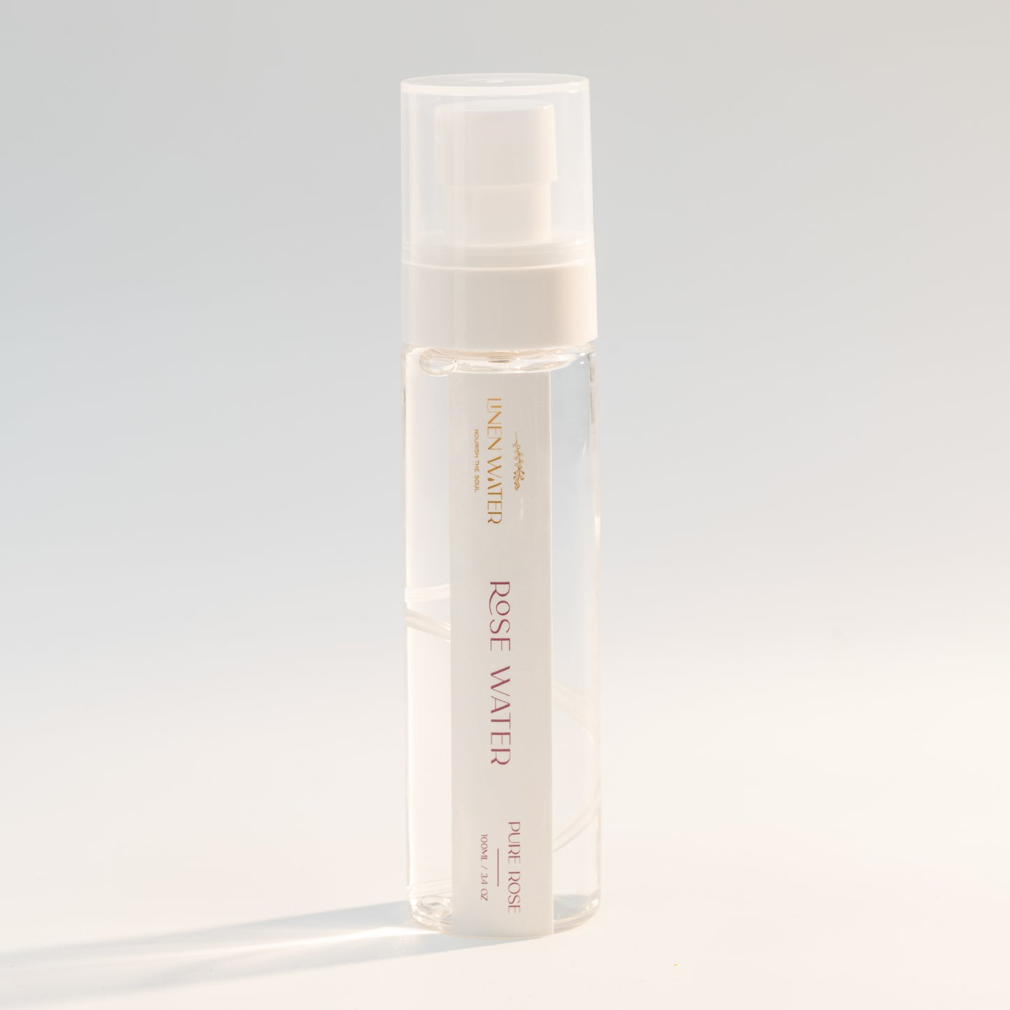 A clear glass spray bottle with the label "Linen Water Rose Water" is captured against a soft white backdrop.  Its basic, refreshing essence for natural skincare is highlighted by the golden details and clean lines.
