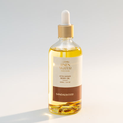 The warm amber tone and stylish packaging of Linen Water Sandalwood Body Oil are shown in a clear glass bottle with a golden dropper against a soft light background.